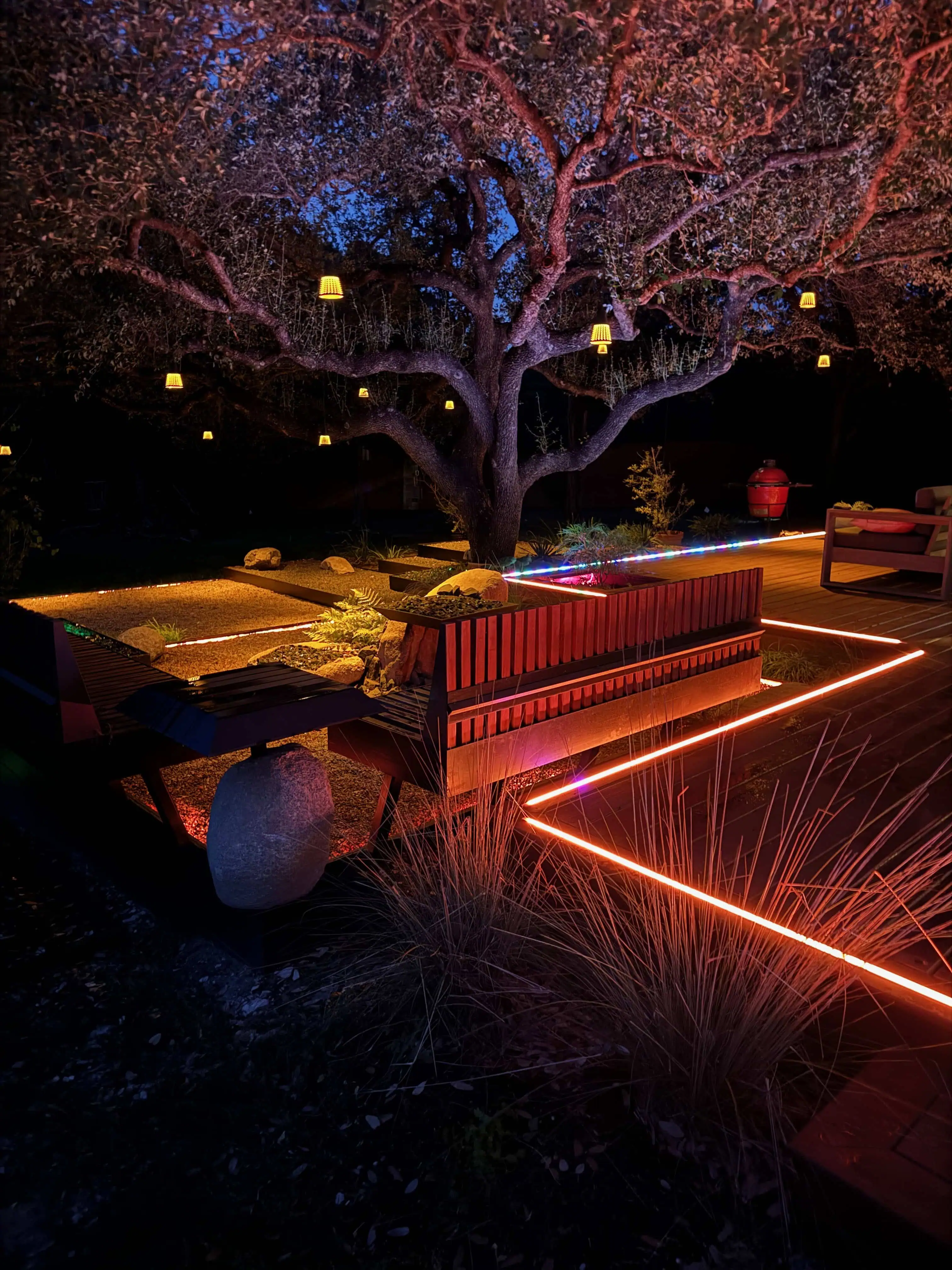 Night view — live oak with hanging lanterns, LED strip lighting along terraced deck and steel edges — Circle C Austin TX